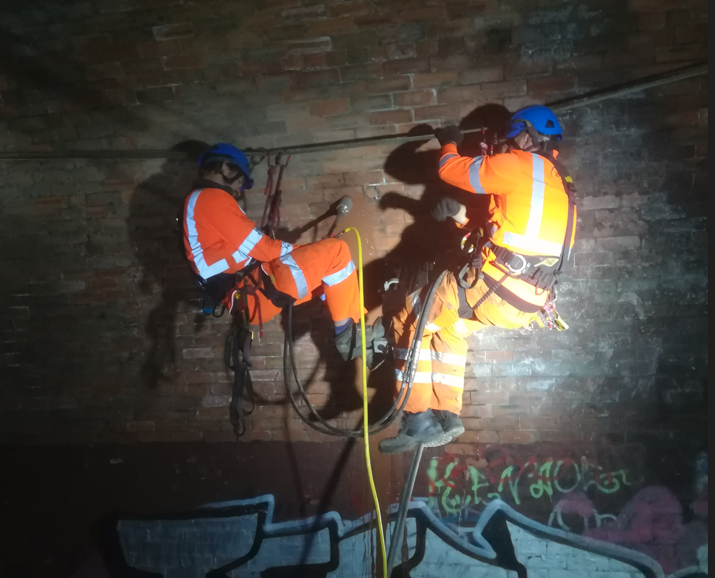 Two rope access operatives suspended against a brick wall inside a dark structure, coring the masonry by torchlight.